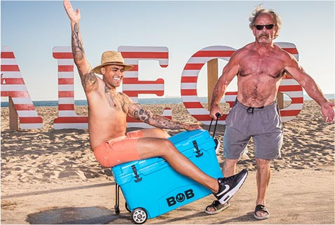 A muscular man pulling a 75QT / 70.97L hard cooler while another man straddles the cooler and rides on top of it.