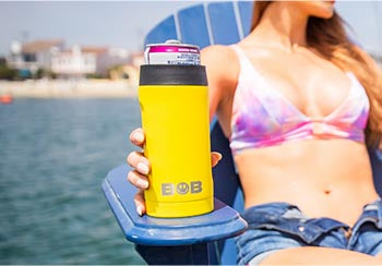 A woman sitting on a chair on a dock and holding a drink in a 12oz Slim “Bob's Chillin” Can Cooler.