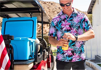 A man opening a drink in a can cooler whiles standing next to a hard cooler on the back of a golf cart.